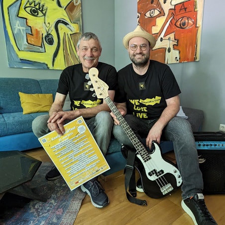 Das Foto zeigt die beiden Organisatoren mit Plakat, Bassgitarre und Verstärker.