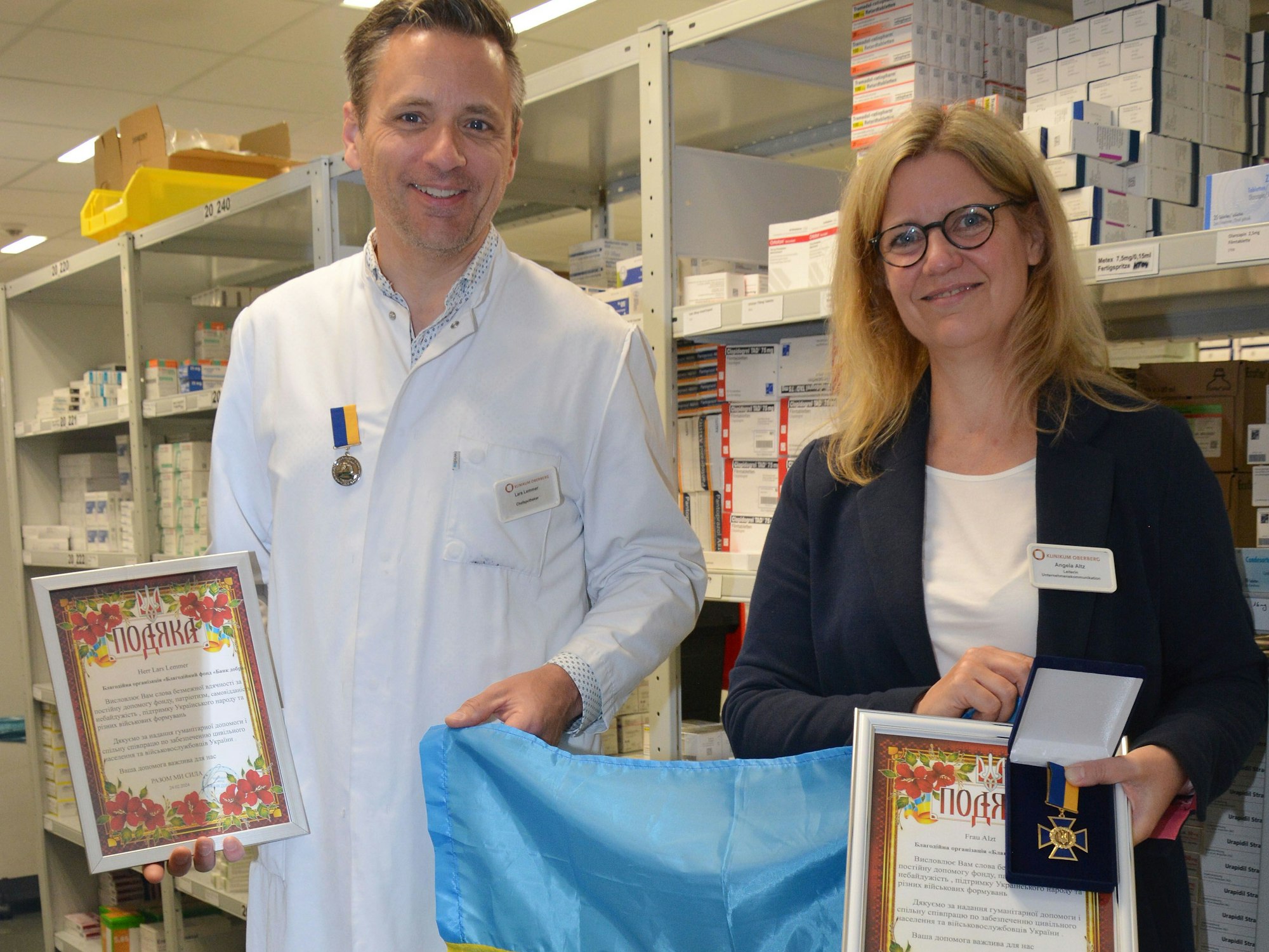 Angela Altz und Lars Lemmer vom Klinikum Oberberg stehen mit einer Ukraine-Flagge, Urkunden und einem Verdienstorden nebeneinander.
