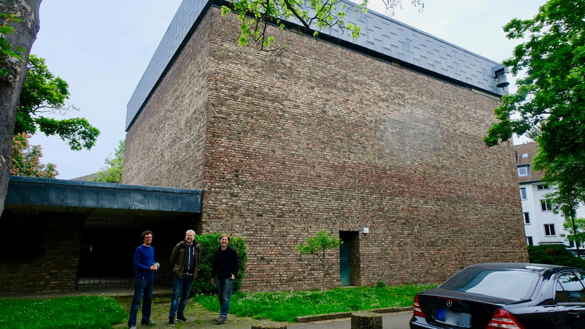 Vor einem hohen würfelförmigen Beton-Gebäude stehen drei Männer.