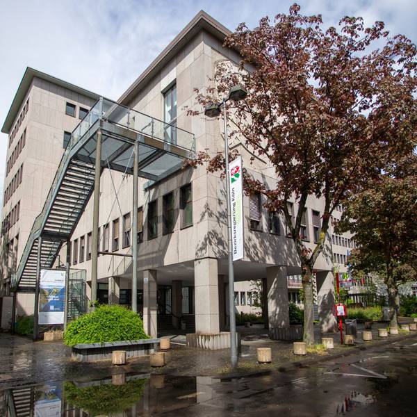 Das Gebäude der Bezirksregierung Köln in der Innenstadt ist zu sehen. Eine moderne Treppenkonstruktion zieht sich vor der Fassade in den dritten Stock. Am Straßenrand stehen Bäume.