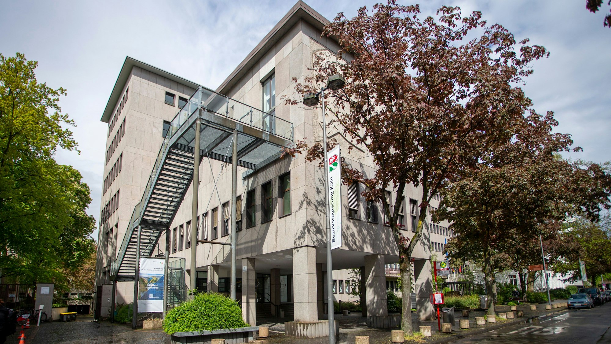 Das Gebäude der Bezirksregierung Köln in der Innenstadt ist zu sehen. Eine moderne Treppenkonstruktion zieht sich vor der Fassade in den dritten Stock. Am Straßenrand stehen Bäume.