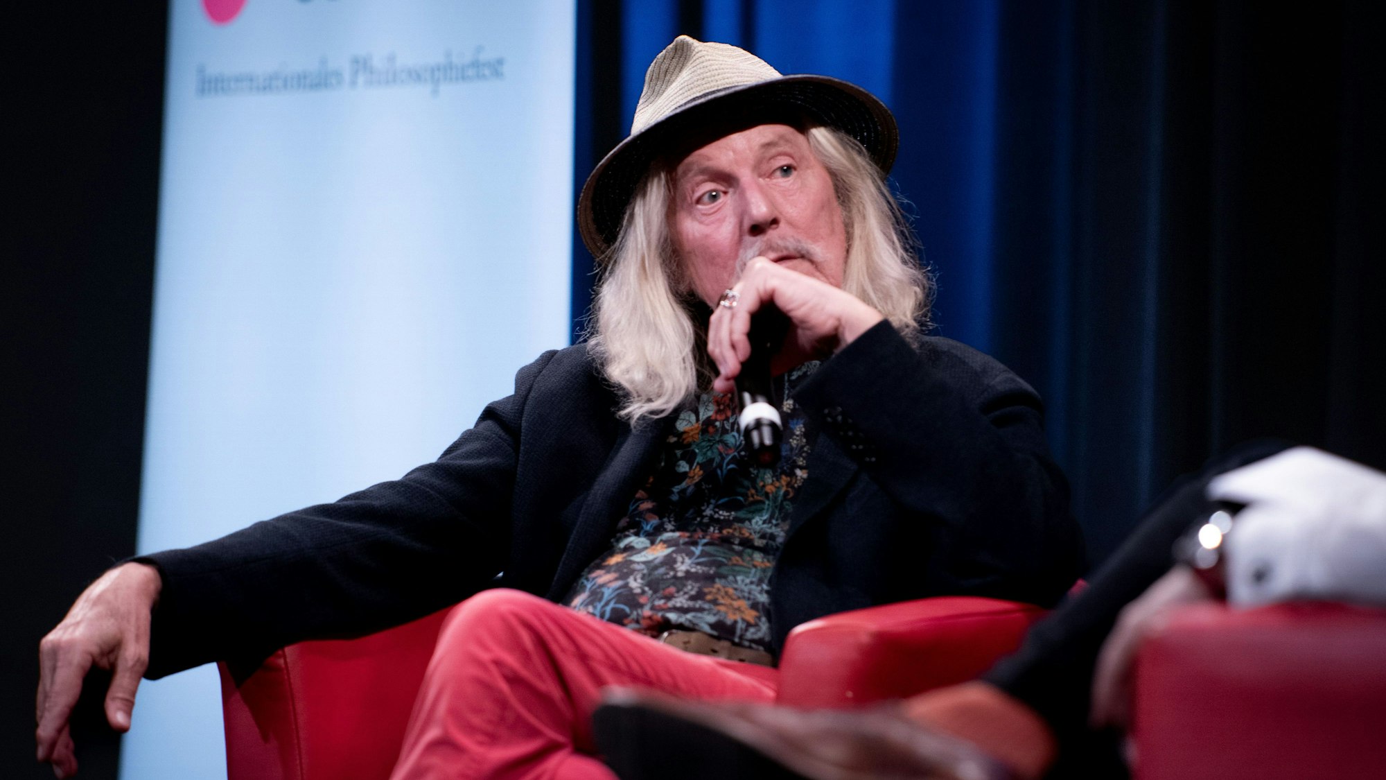 Wolfgang Beltracchi sitzt auf einem roten Sessel und überkreuzt die Beine. Er trägt ein buntes Hemd und einen Hut. Er hat schulterlange Haare.