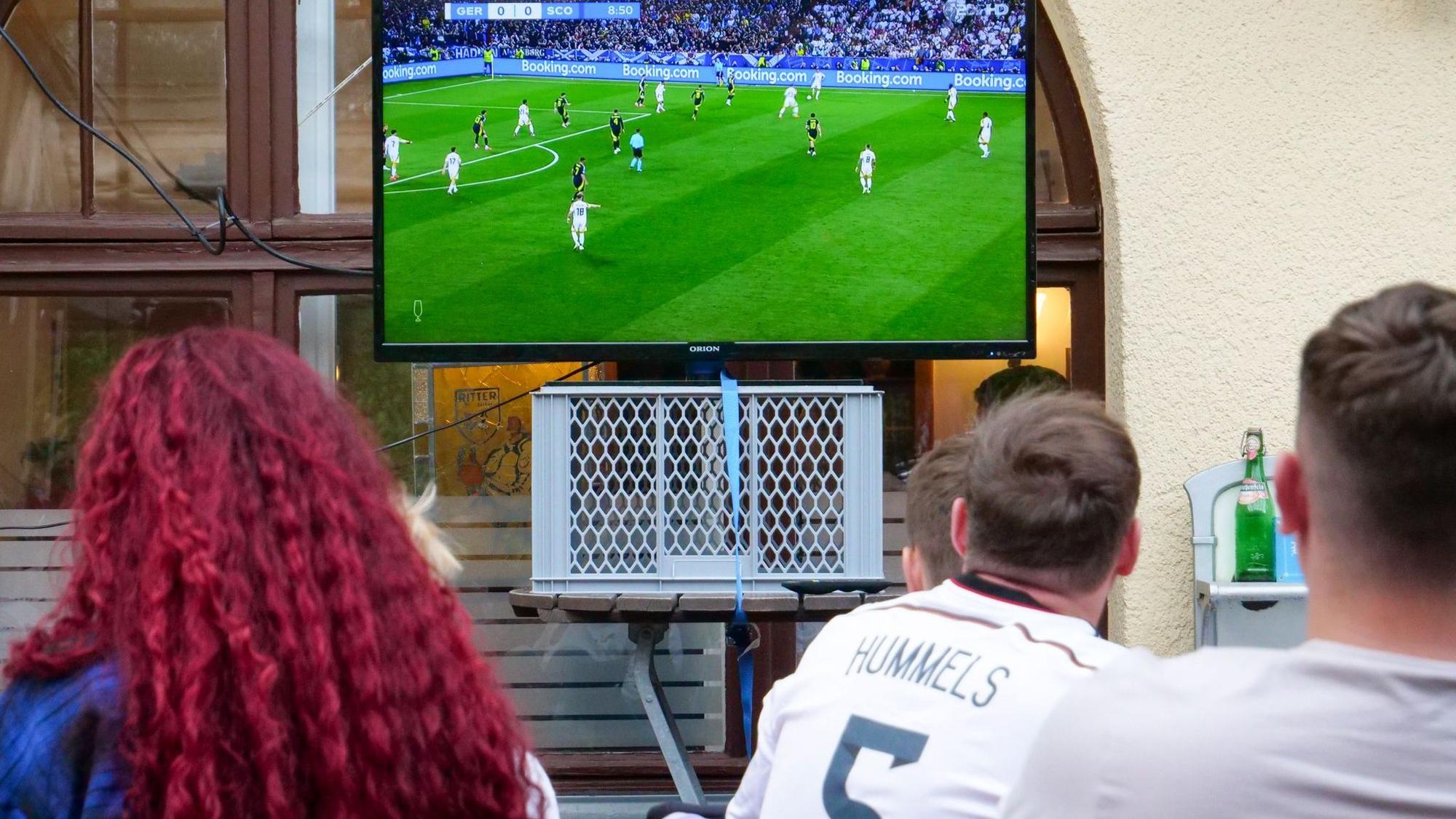Auf dem Foto sind Fußball-Fans zu sehen, die das Spiel auf einem Fernseher im Biergarten verfolgen.