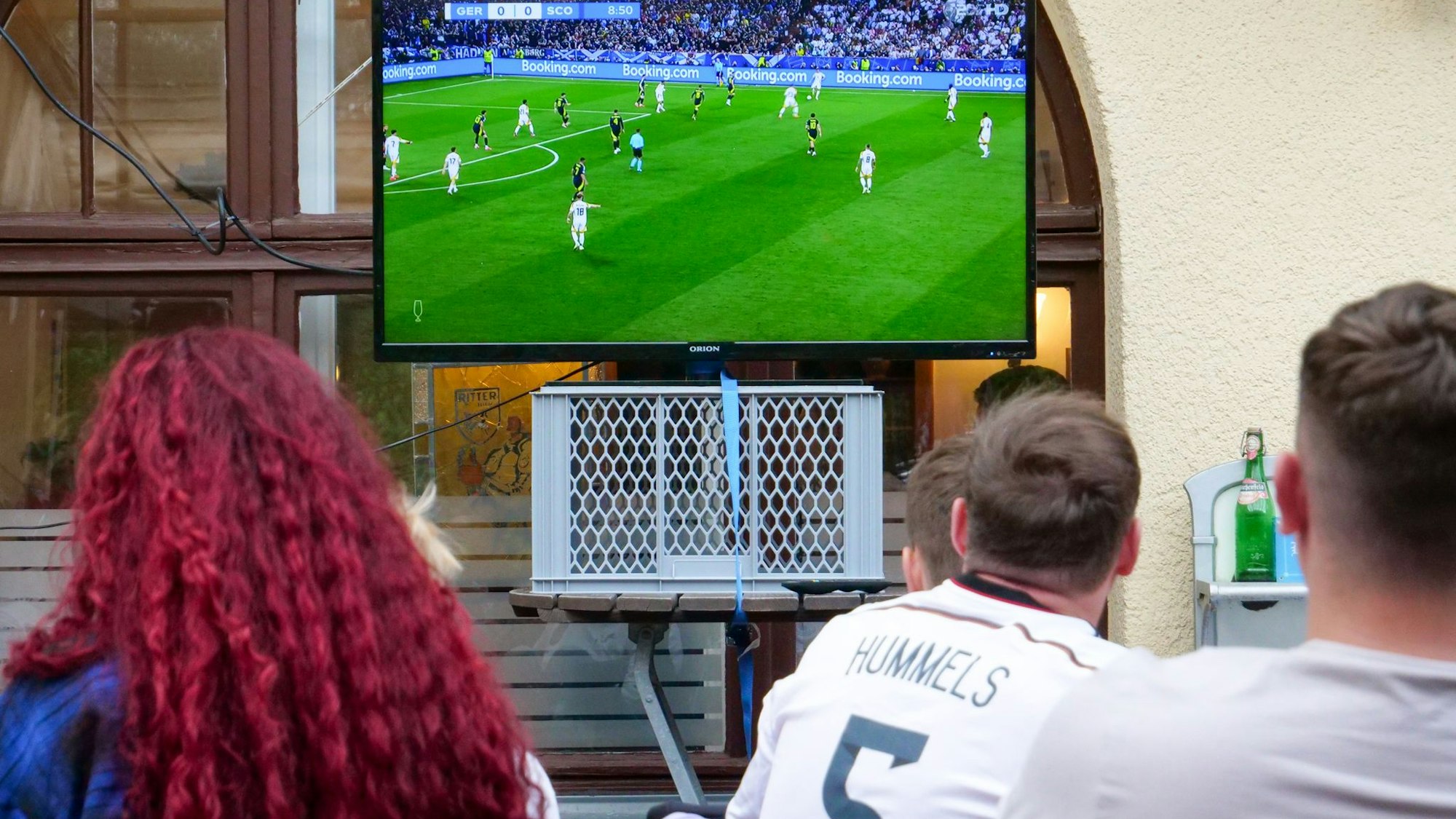 Auf dem Foto sind Fußball-Fans zu sehen, die das Spiel auf einem Fernseher im Biergarten verfolgen.