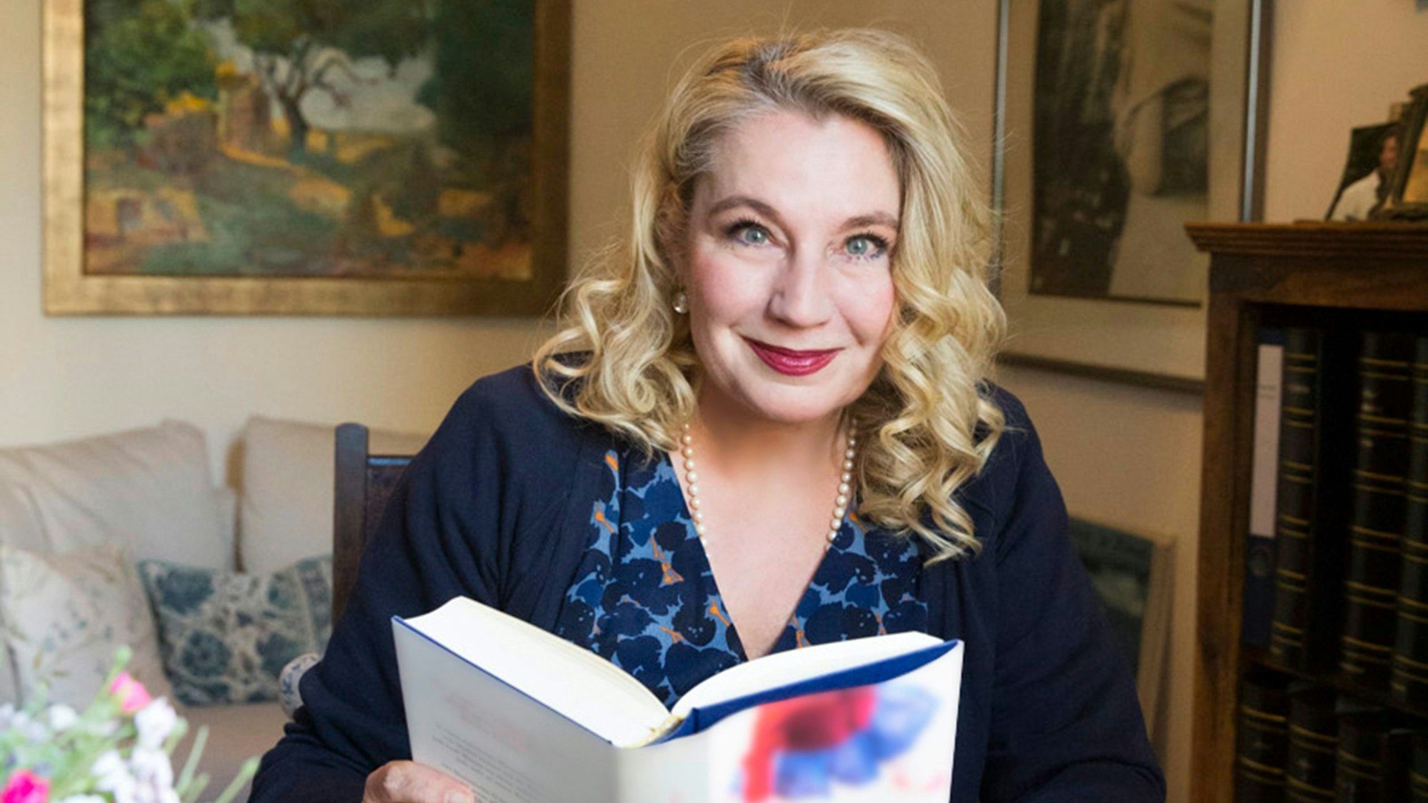 Eine Frau mit blonden Haaren sitzt mit einem Buch an einem Tisch.