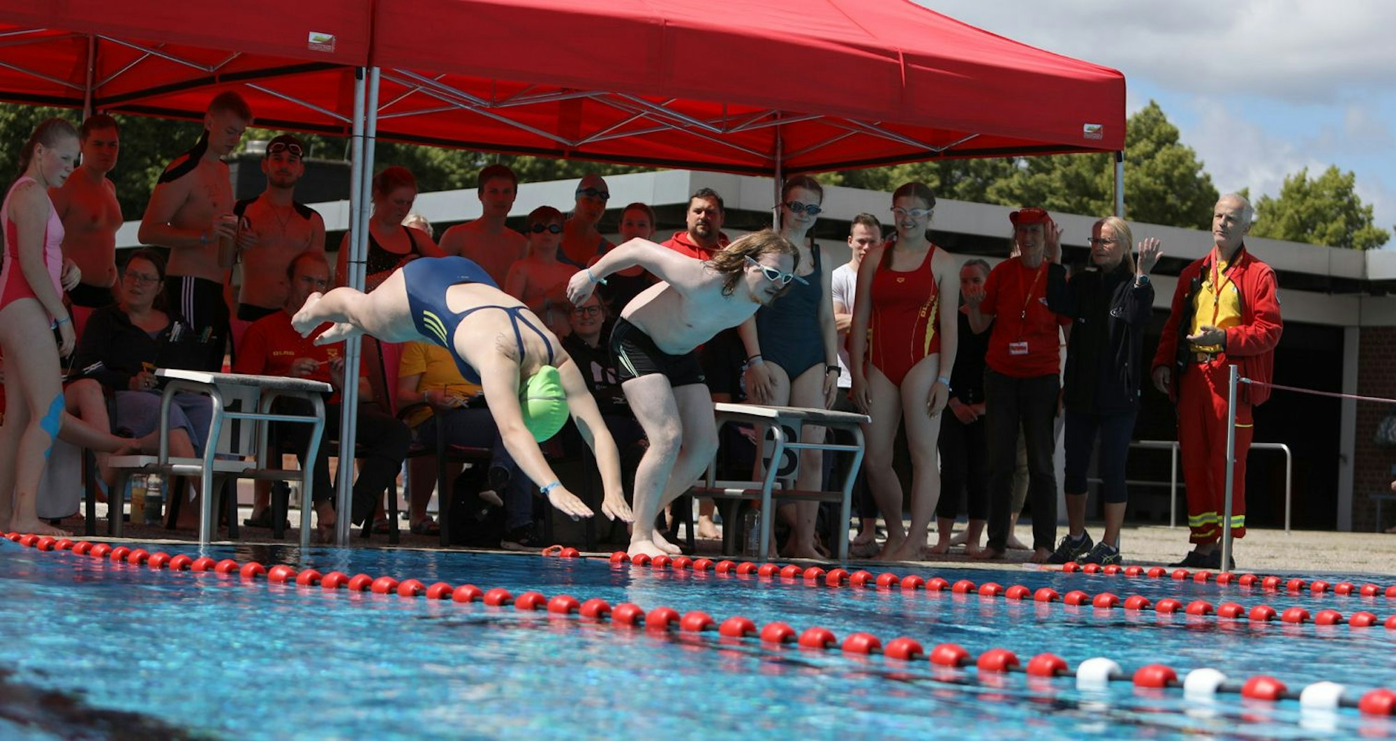 Zwei Menschen springen mit einem Kopfsprung in ein Schwimmbecken, hinter ihnen stehen zahlreiche Zuschauer.