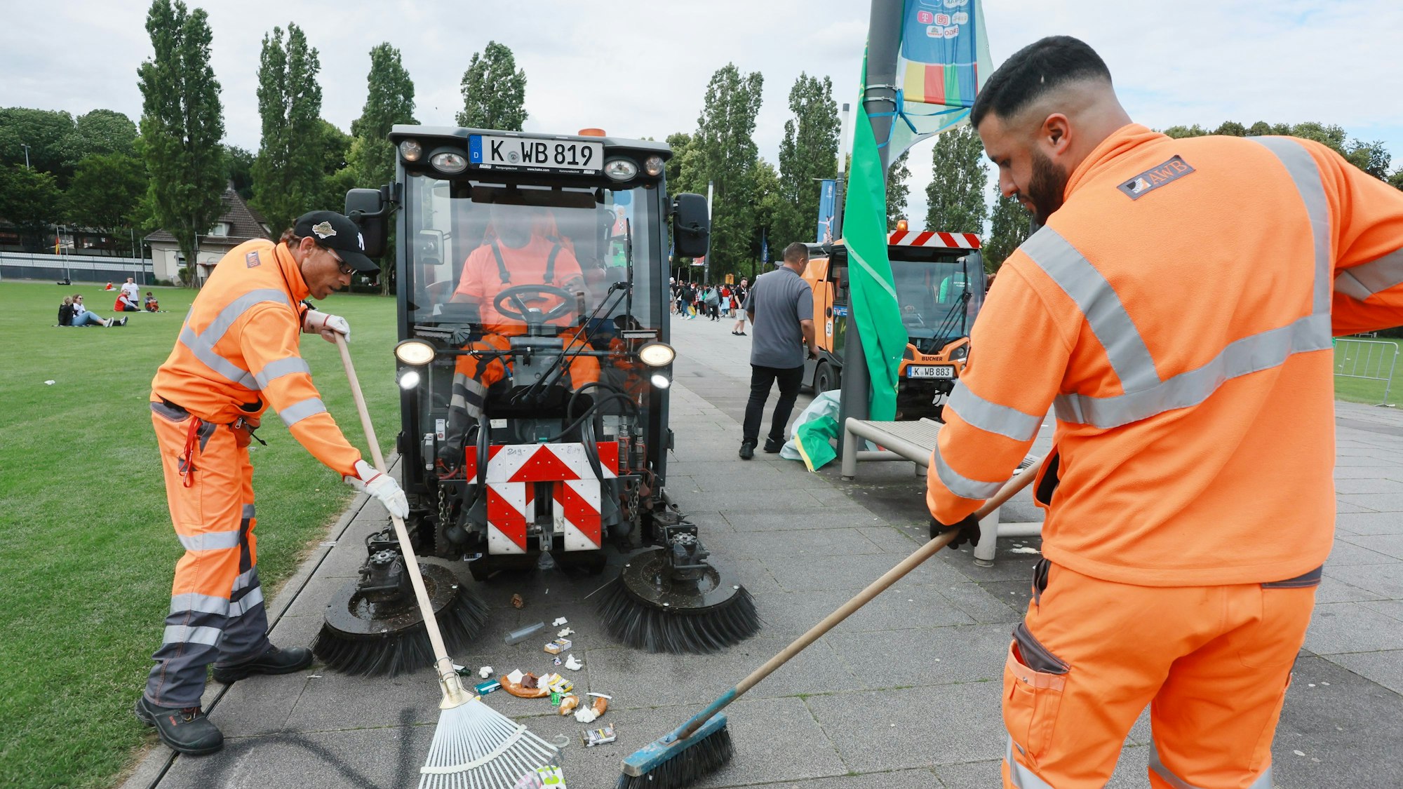 Mitarbeitende der AWB Köln kehren Müll vor dem Weg am Rhein-Energie-Stadion zusammen. Dabei helfen ihnen Kehrmaschinen.