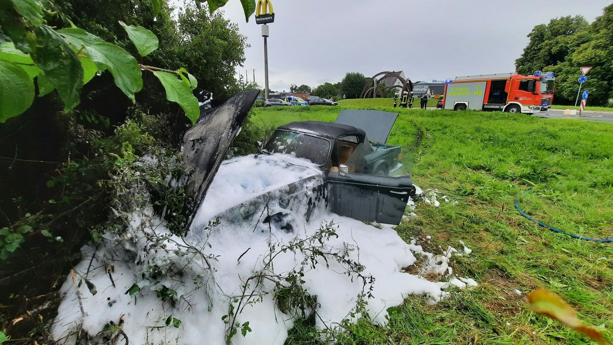 Das mit Löschschaum bedeckte Wrack eines ausgebrannten Oldtimers steht auf einer Wiese nahe der Autobahnauffahrt Siebengebirge.