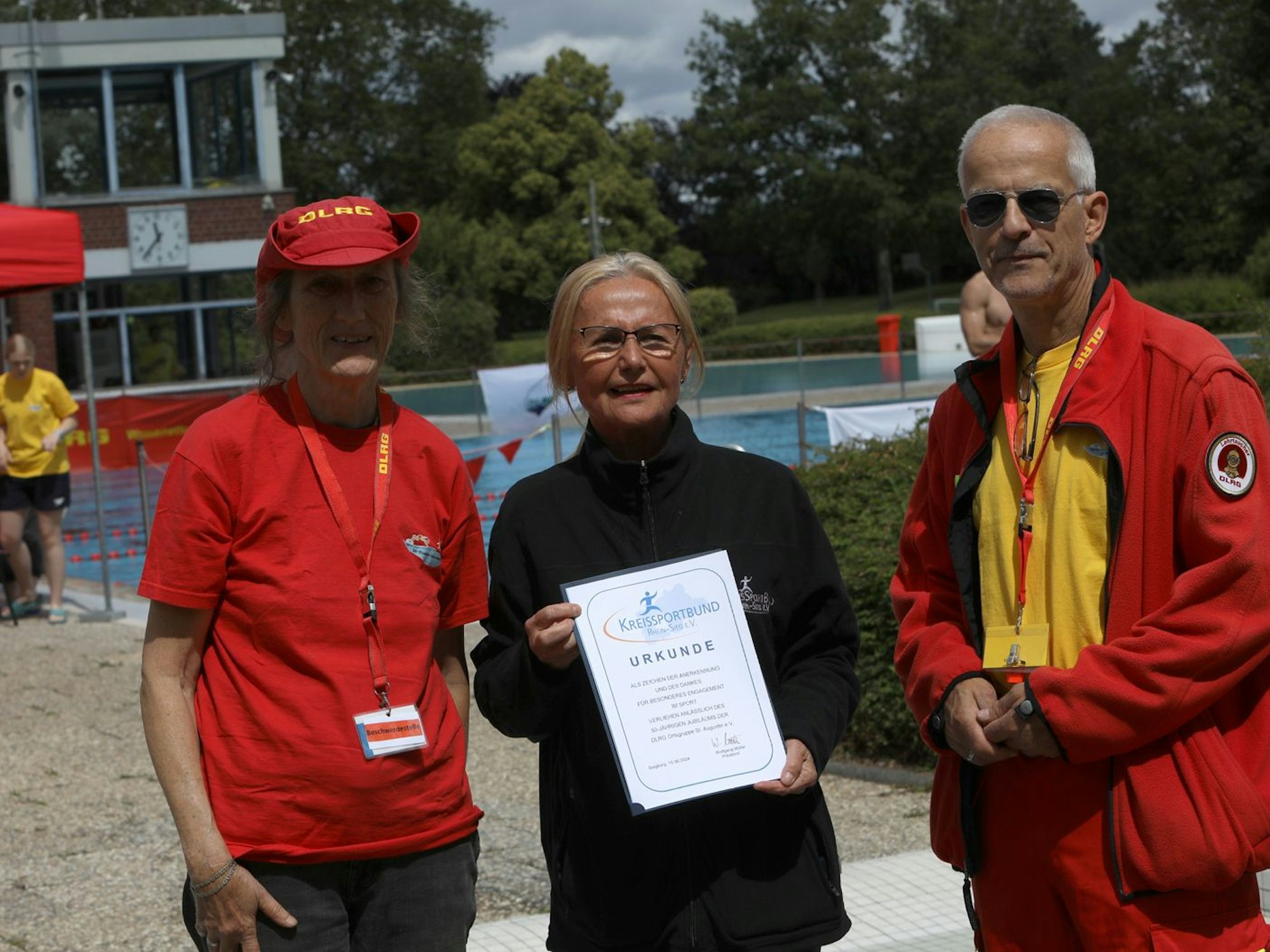 Eine Frau im schwarzen Pullover hält ein Papier mit der Aufschrift "Urkunde" vor sich, links und rechts neben ihr stehen eine Frau und ein Mann, beide tragen rote Kleidung.