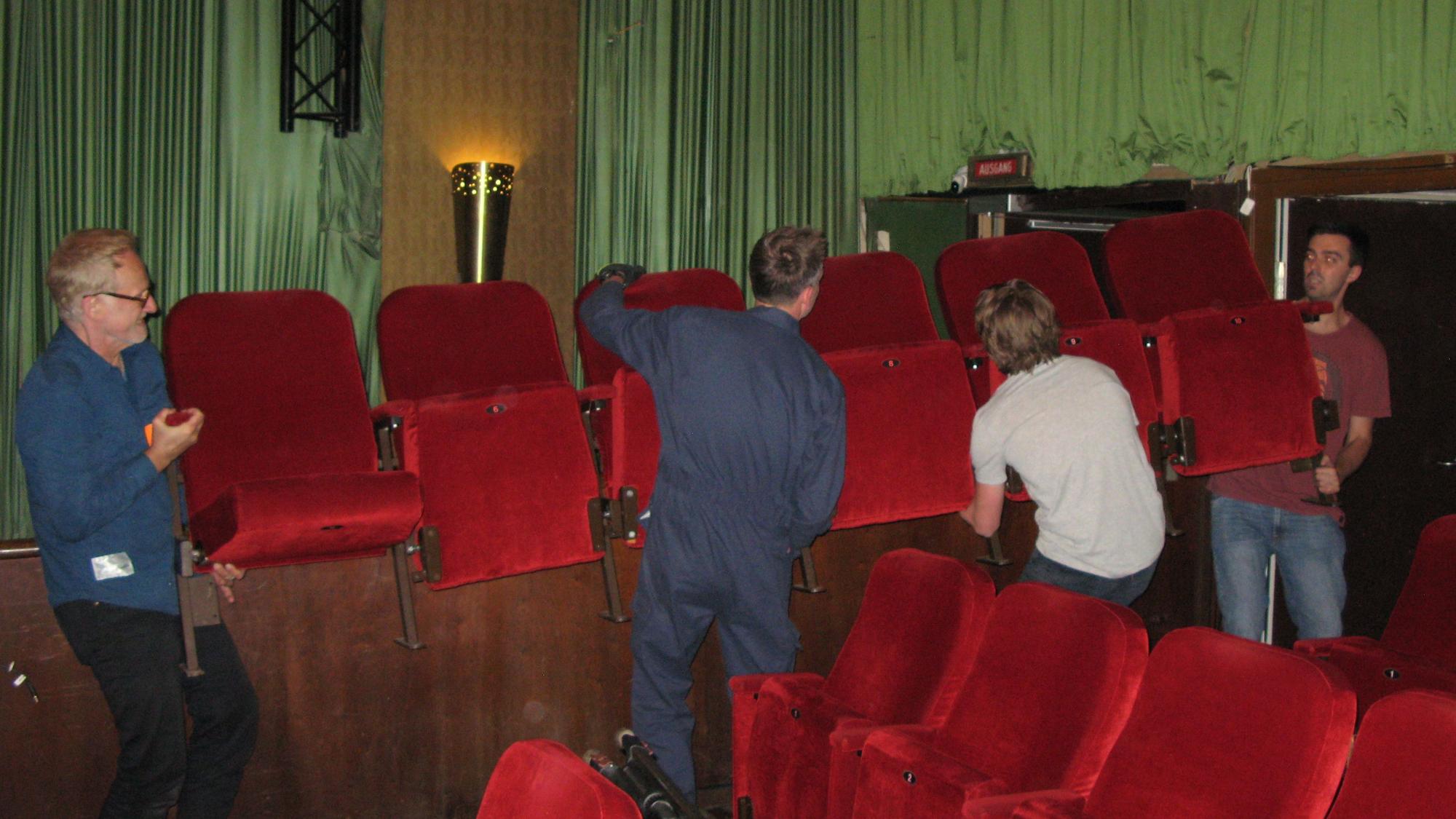 Vier Männer schleppen eine rote Kino-Klappsesselreihe aus dem Kinosaal.