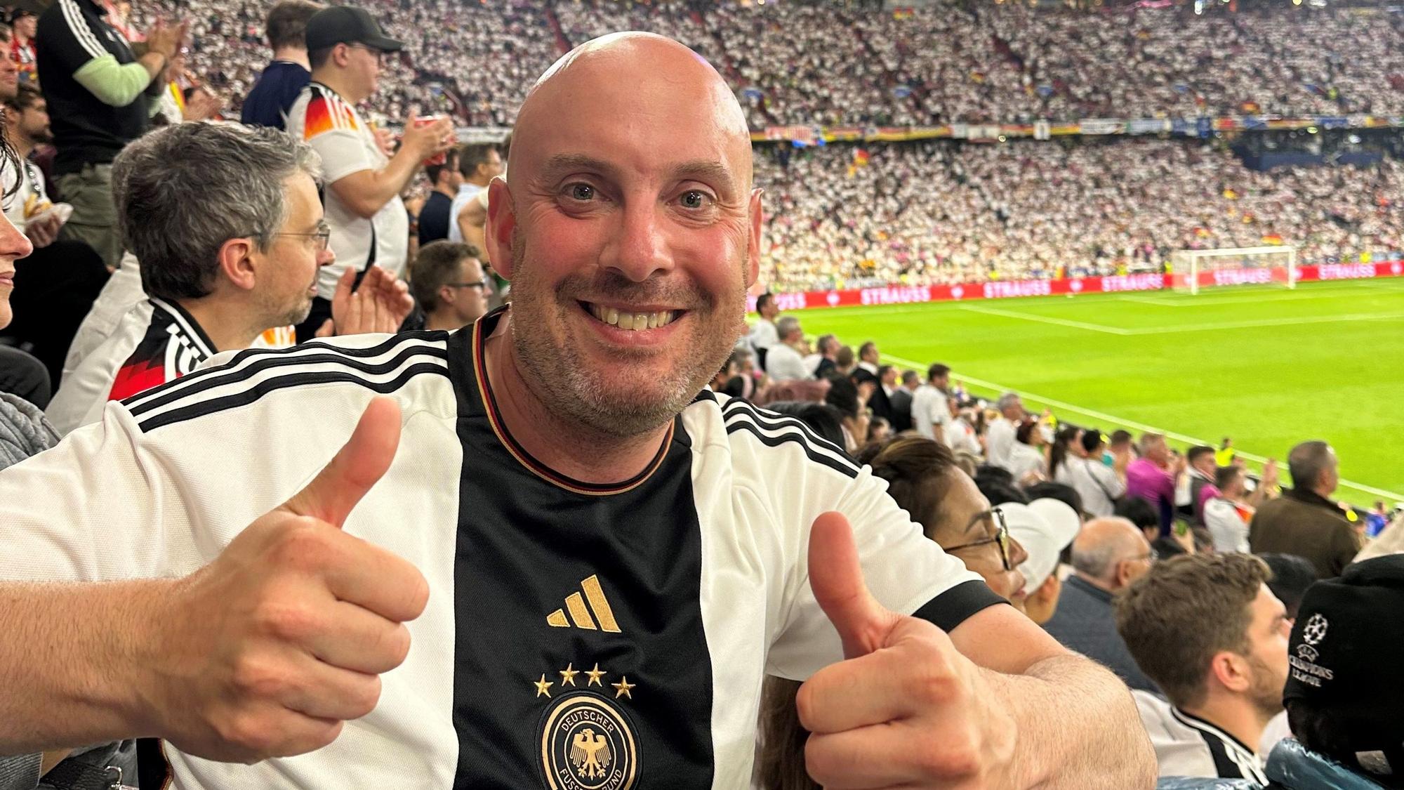 Andre Knips schaut in die Kamera und hebt die Daumen. Im Hintergrund sieht man die vollbesetzte Münchener Arena.