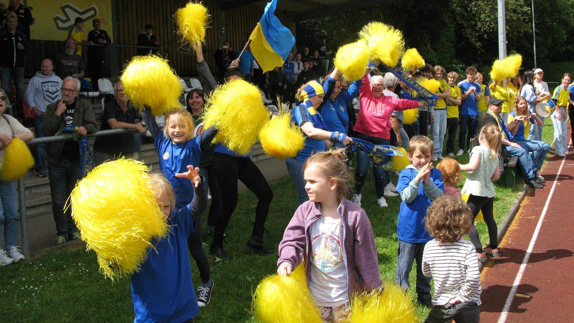 Zuschauer, Erwachsene und Kinder, wedeln fröhlich mit gelben Pompons.