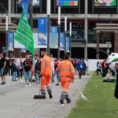Zwei Mitarbeiter der AWB laufen zwischen Fußballfans umher und kehren Müll auf dem Weg vor dem Stadion zusammen.