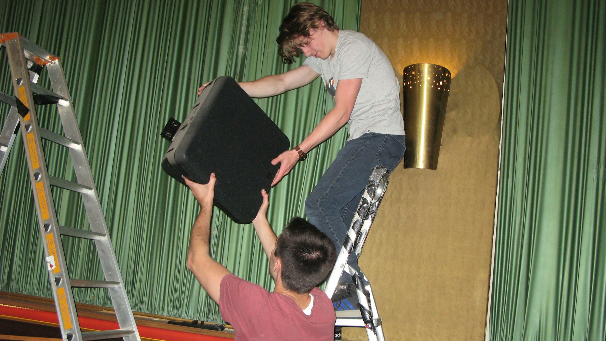 Zwei junge Männer mit Leitern, der eine reicht dem anderen einen demontieren Lautsprecher.