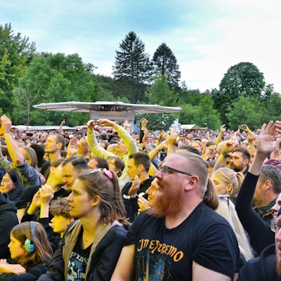 Das Bild zeigt einen Teil der Zuschauer, die stehend das Konzert verfolgen. Einige tragen T-Shirts mit dem Namen der Band.