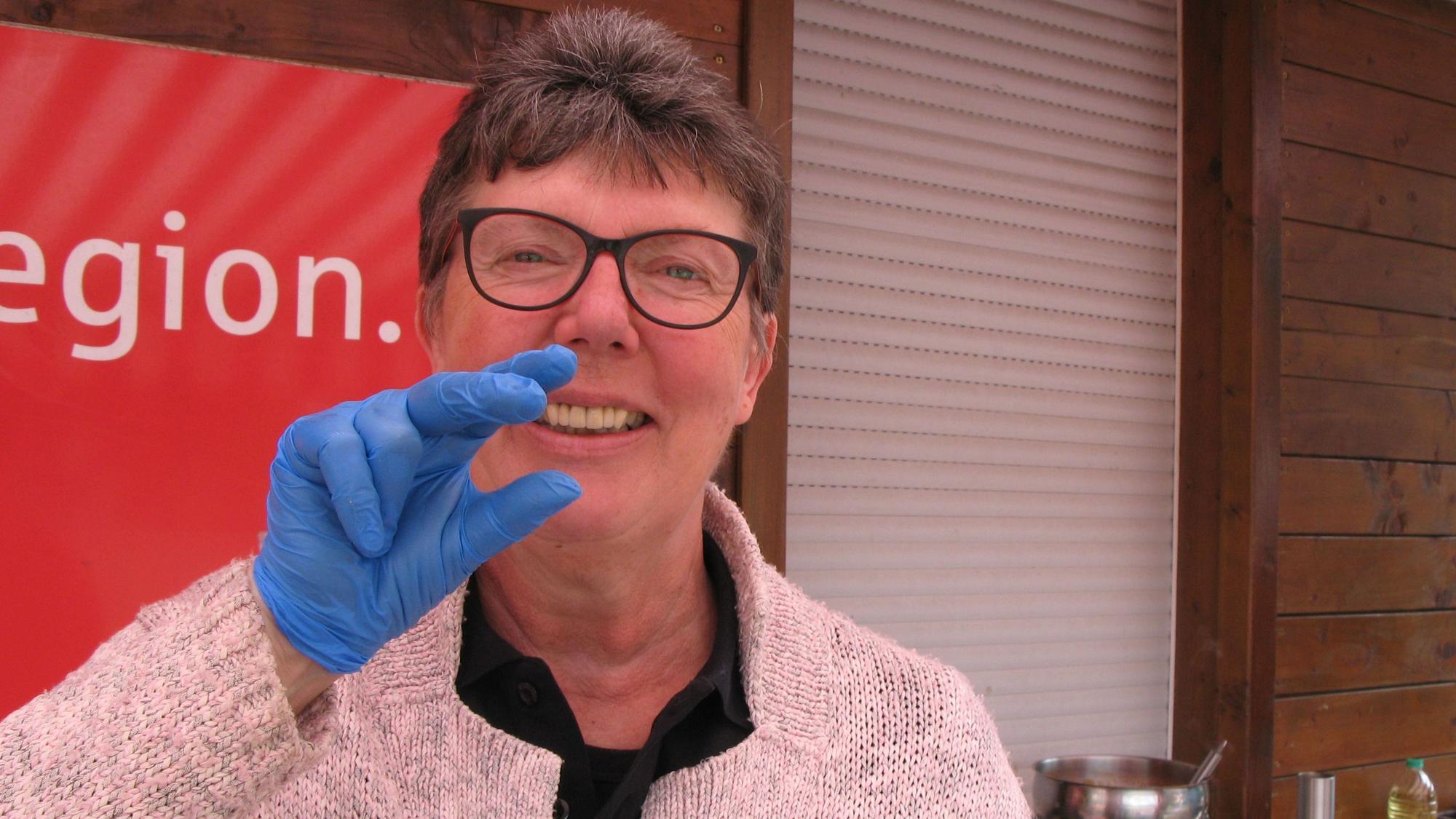 Beate Duderstadt am Grillstand macht die Gebärde für Geflügelwürstchen: die Hand als Schnabel vor dem Mund.