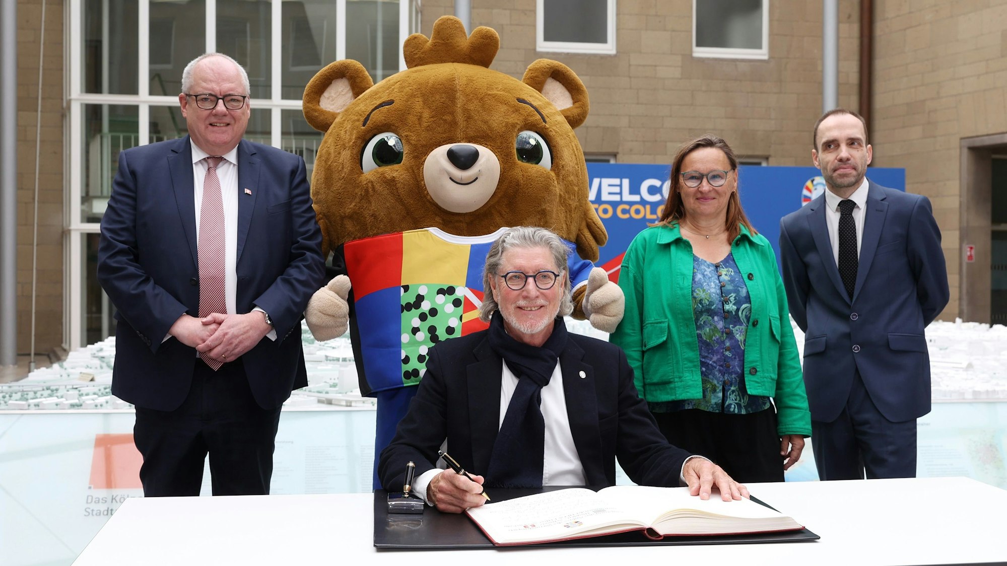 Vier Menschen stehen bzw. sitzen an einem Tisch, hinter ihnen steht das EM-Maskottchen.