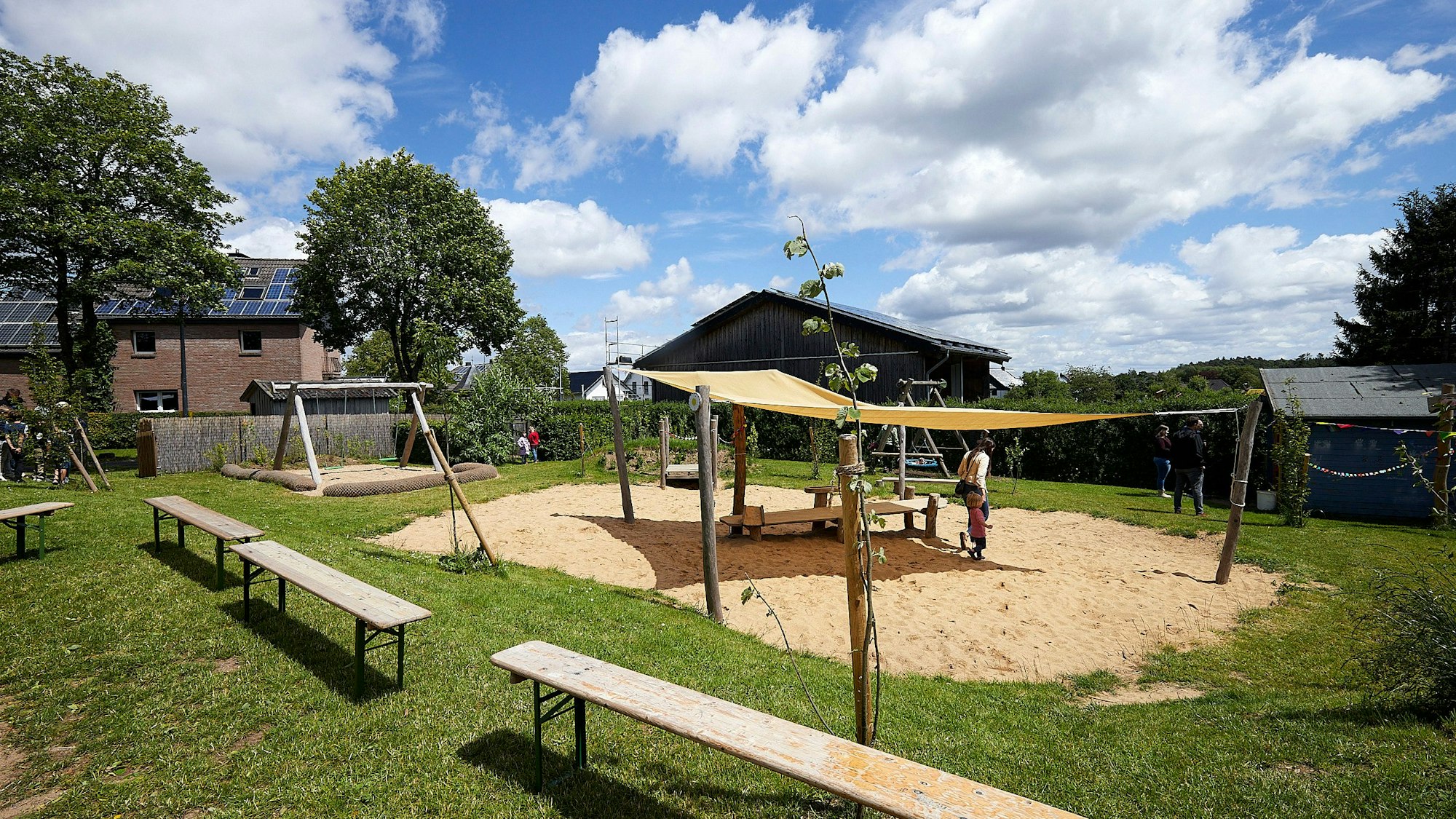 Das Außengelände mit Spielgeräten, Bäumen und einer überdachten Sandfläche.