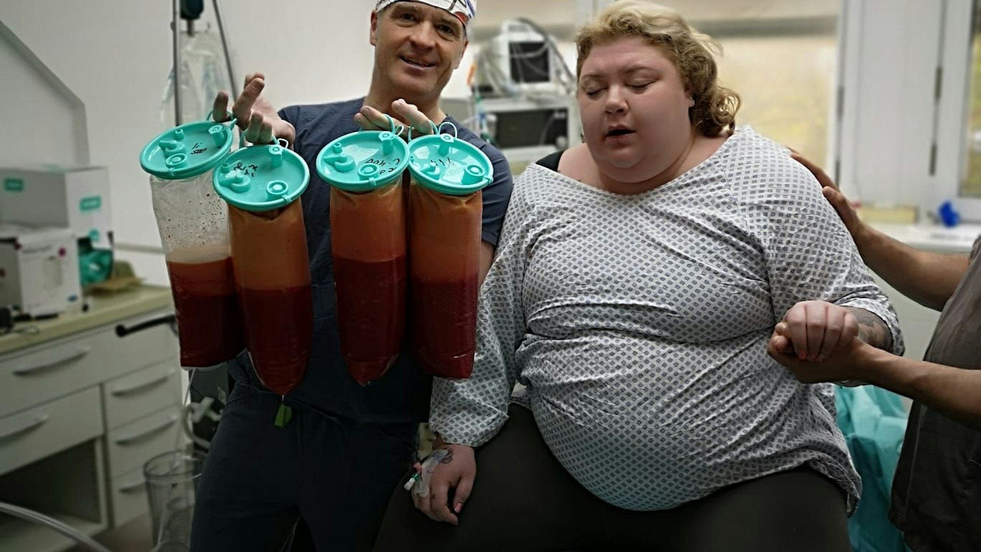 Sandra Fritzler sitzt nach einer Operation auf einer Liege. Ihr Operateur hält die Behältnisse in die Luft, in denen sich Fett befindet.