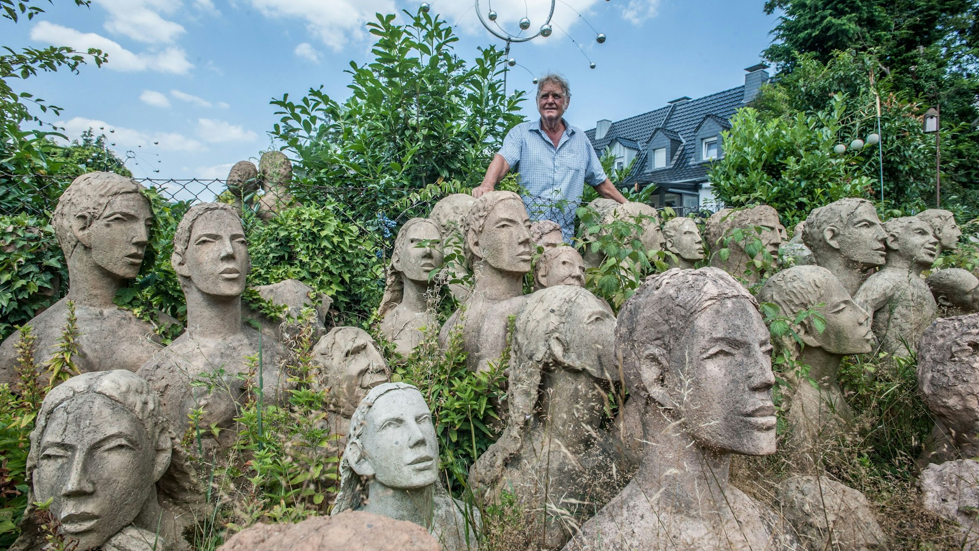 Der Künstler Richard Bausewein steht in seinem Garten, den er zeitgleich als Atelier nutzt, vor einer Vielzahl an Skulpturen, die nur aus Oberkörpern bestehen.