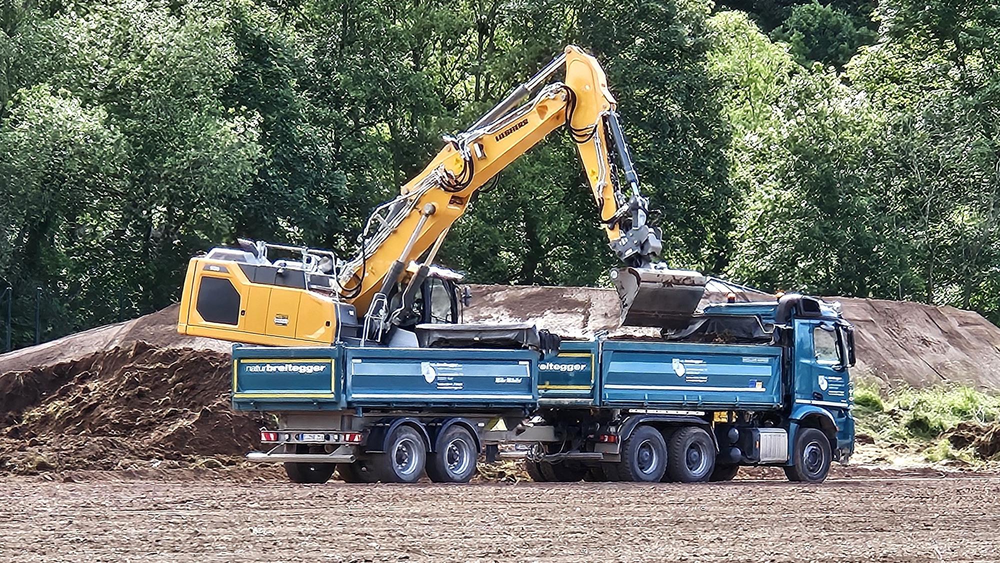 Ein Bagger belädt einen Lkw mit Anhänger mit Erde.
