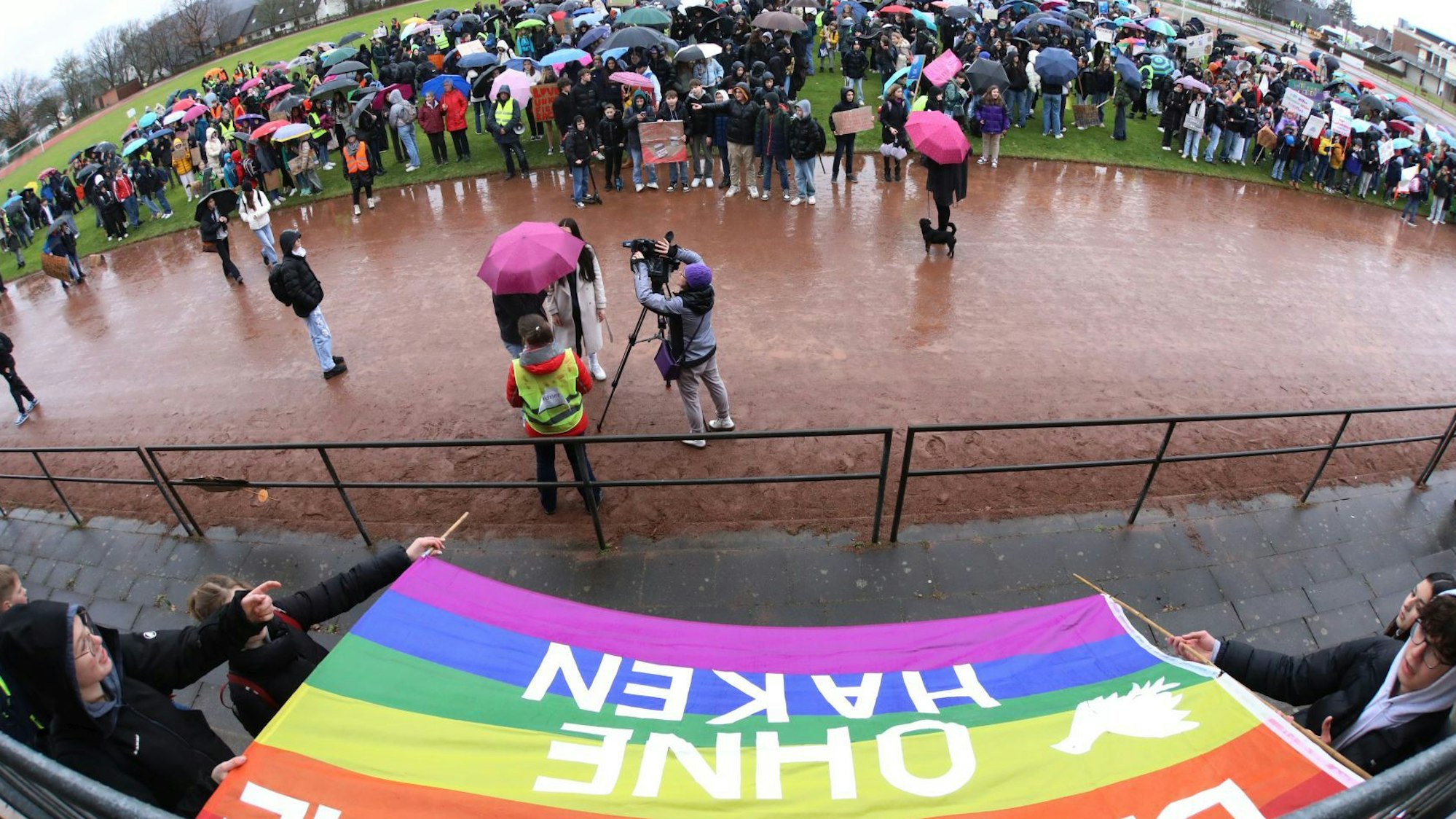 Menschen stehen unter Regenschirmen auf einem Sportplatz.