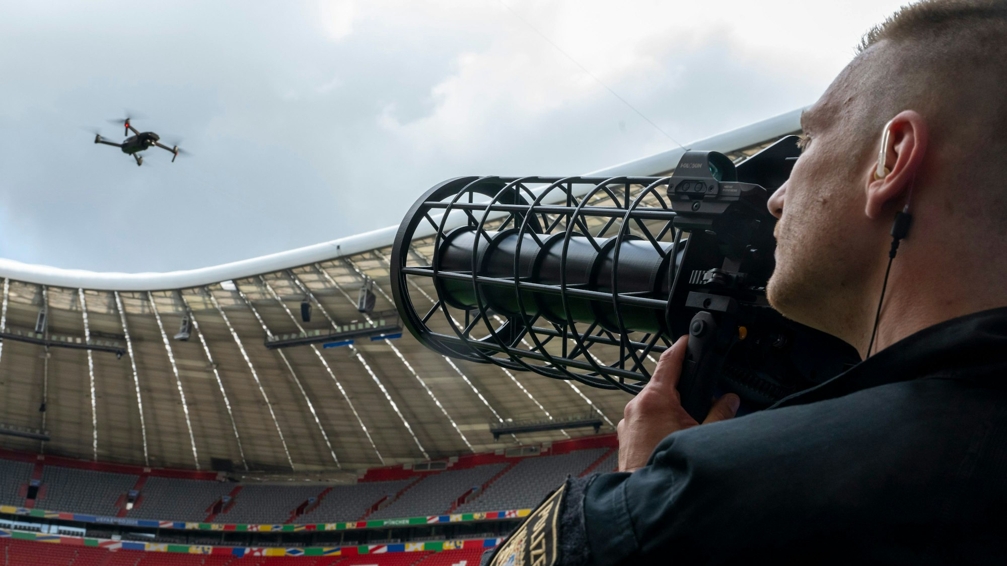 Ein speziell ausgebildeter Polizist zeigt im Fußballstadion München mit einem schwarzen länglichen Gerät gen Himmel, wo eine Drohne fliegt.