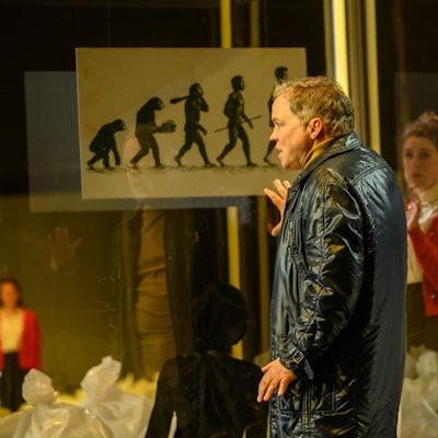 Auf dem Bild trägt Hagen Matzeit eine schwarze Lederjacke. Er lehnt die Hand an eine Scheibe, auf der die Evolution des Menschen dargestellt ist.