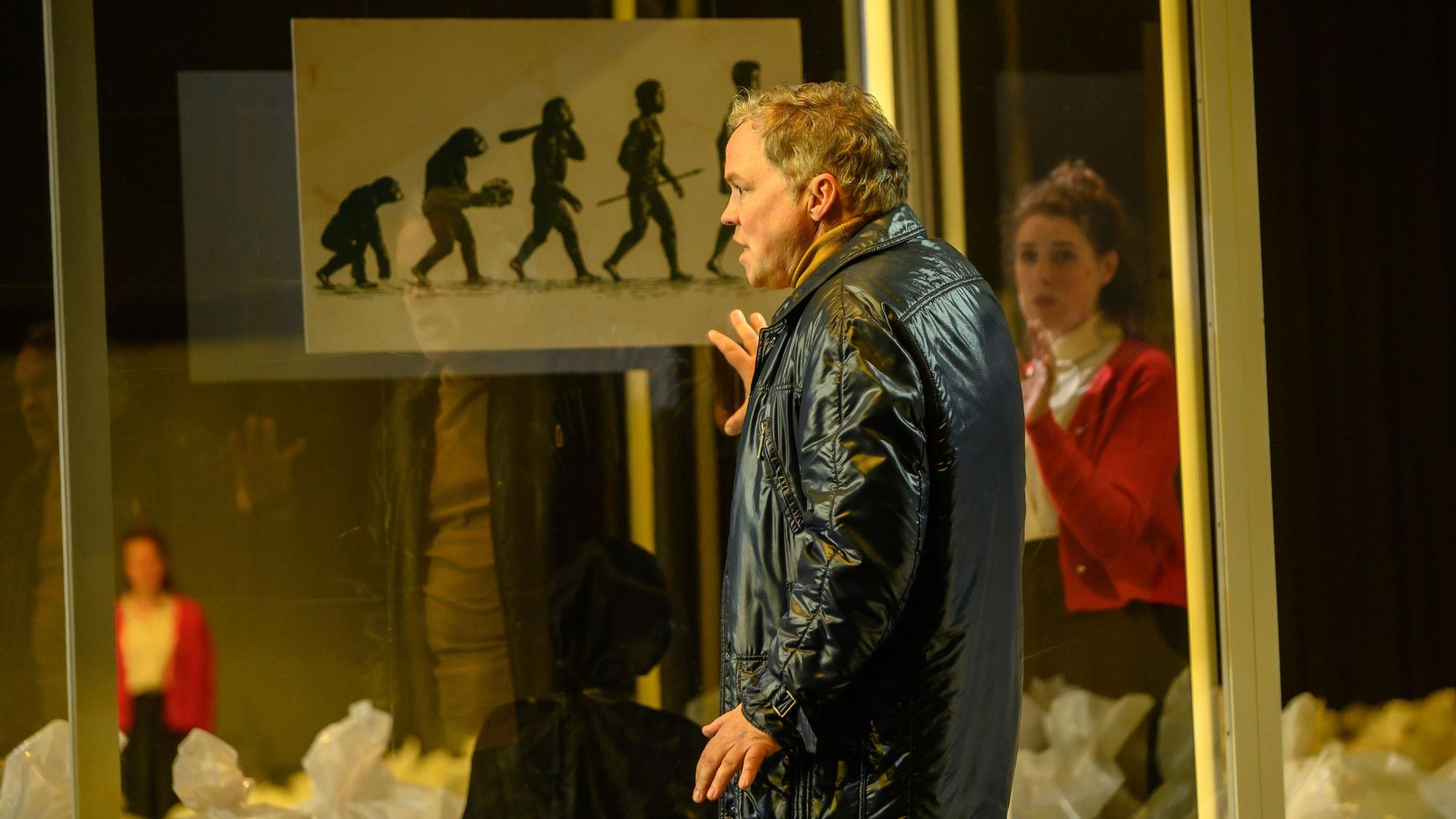 Auf dem Bild trägt Hagen Matzeit eine schwarze Lederjacke. Er lehnt die Hand an eine Scheibe, auf der die Evolution des Menschen dargestellt ist.