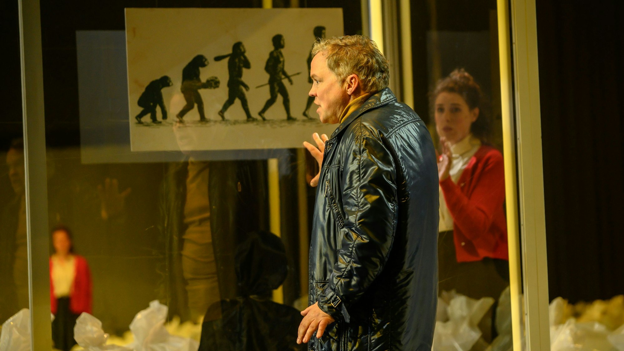 Auf dem Bild trägt Hagen Matzeit eine schwarze Lederjacke. Er lehnt die Hand an eine Scheibe, auf der die Evolution des Menschen dargestellt ist.