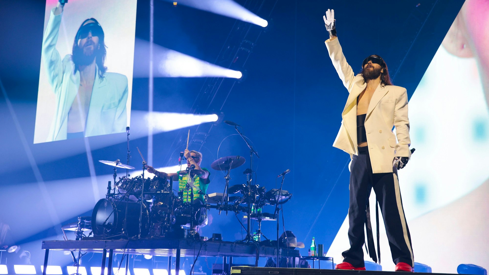 Leadsänger Jared Leto steht im Vordergrund, sein Bruder und Schlagzeuger Shannon sitzt links von ihm an seinem Gerät. Im Hintergrund zeigen Bildschirme Jared in Großaufnahme und das Musikvideo des Songs.