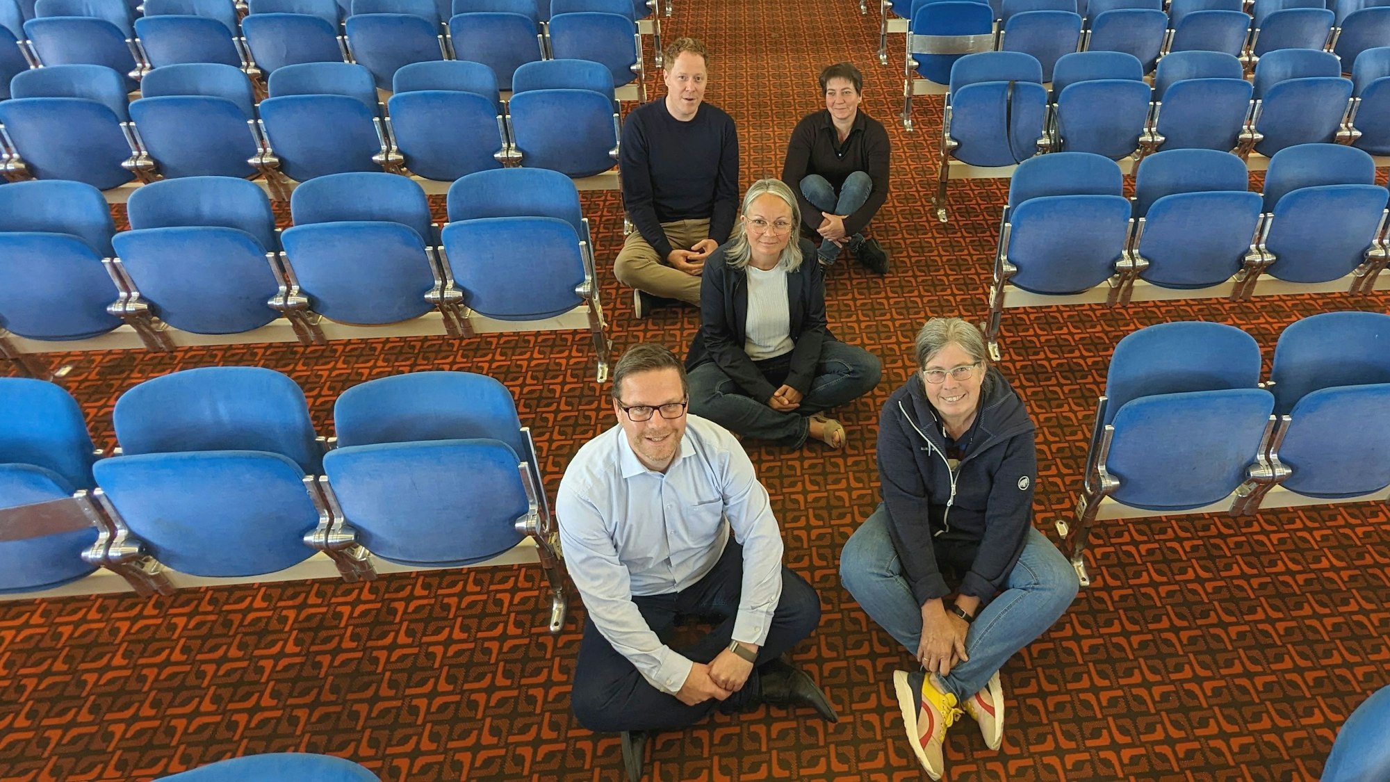 Schulleiter Micha Kreitz und Stellvertreterin Rosemarie Antwerpen (v.l.) sowie Mitglieder des Organisations-Komitees sitzen auf dem Boden der Schulaula.