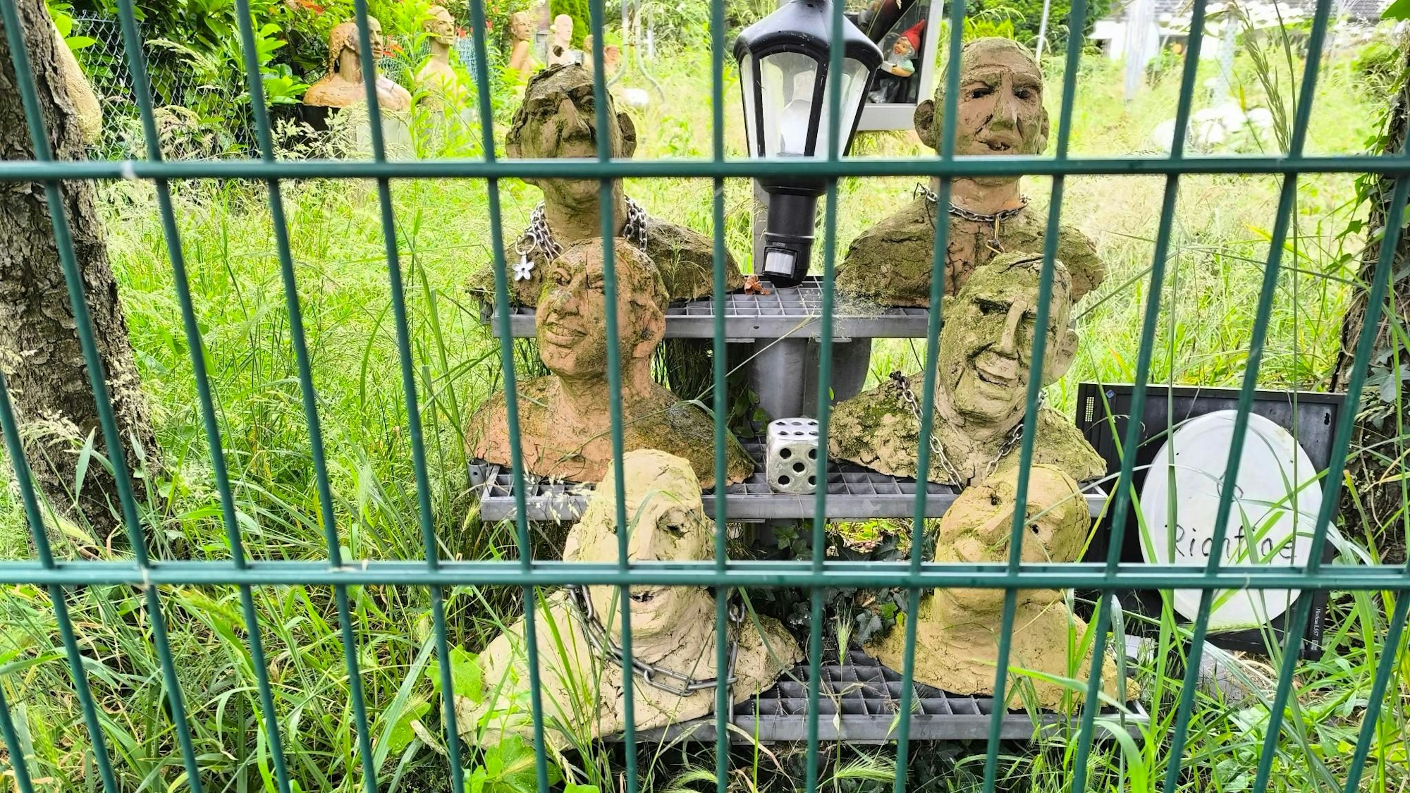 „6 Richtige“ steht neben sechs Skulpturen geschrieben, die auf einer kleinen Treppe hinter einem Gartenzaun zu sehen sind. Um sie herum ist hohes Gras, eine Laterne und ein Würfel sind mittig zwischen den Skulpturen platziert.