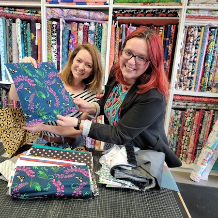 Zwei Frauen lächeln in die Kamera, sie halten Stoffproben in der Hand, im Hintergrund ein Regal mit unendlich vielen Stoffen.