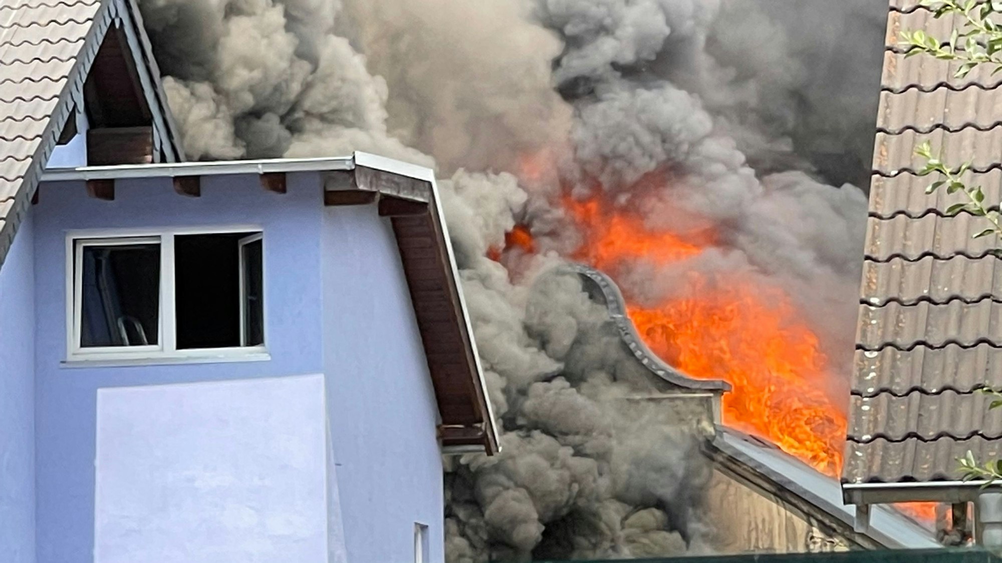 Aus einem Dach quellen schwarze Rauchwolken, rote Flammen schlagen aus der Dachhaut.