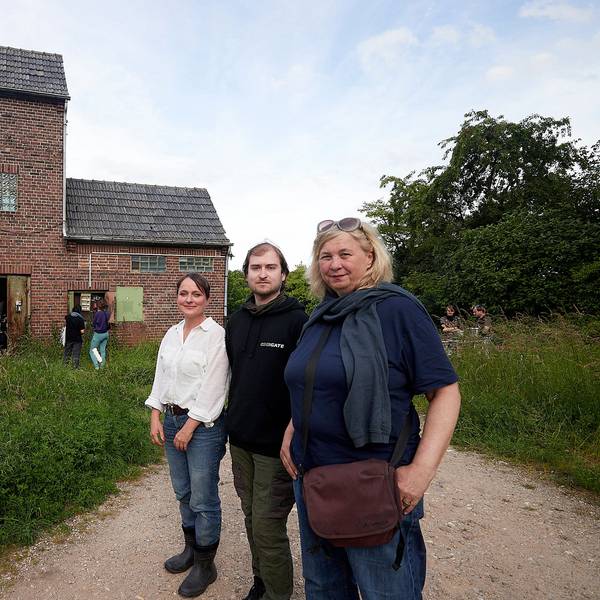 Zwei Frauen und ein Mann, Stefanie Klingemann, Pascal Dreier und Ute Hörner, stehen vor dem alten Trafohaus in Satzvey, das von vielen grünen Sträuchern und Bäumen umgeben ist.