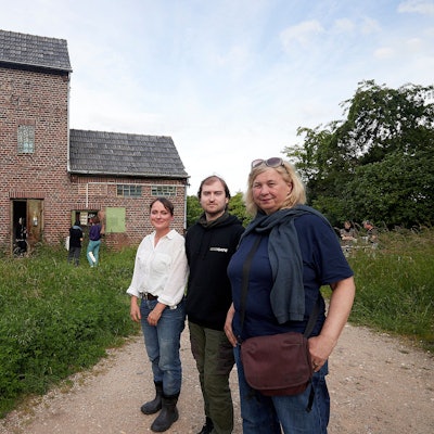Zwei Frauen und ein Mann, Stefanie Klingemann, Pascal Dreier und Ute Hörner, stehen vor dem alten Trafohaus in Satzvey, das von vielen grünen Sträuchern und Bäumen umgeben ist.