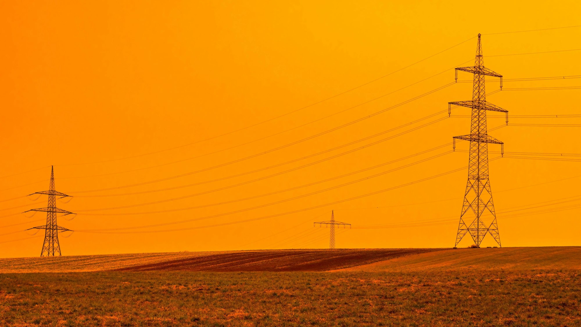 Strommasten stehen auf einem Feld, während der Himmel gelb leuchtet.