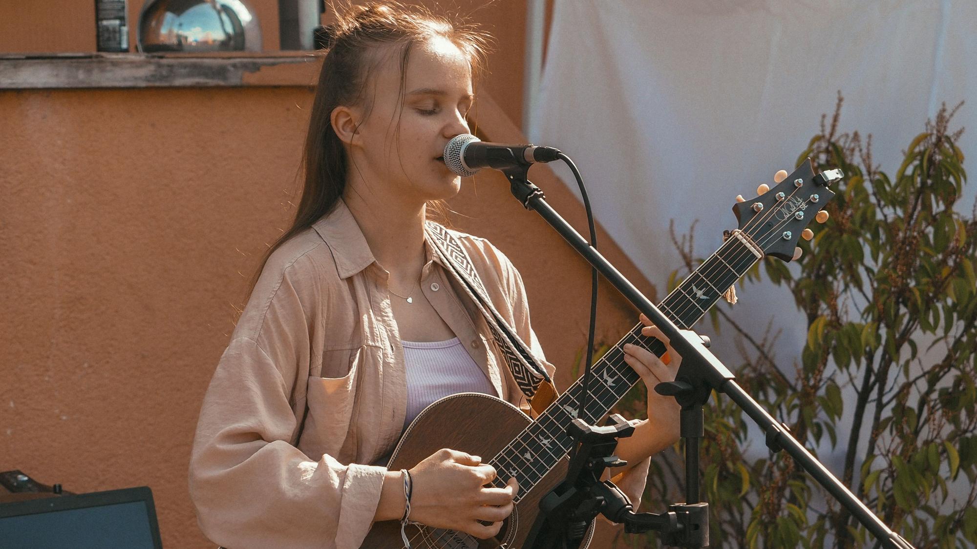 Kira Grohs steht mit einer Gitarre in der Hand vor einem Mikrofon und singt.