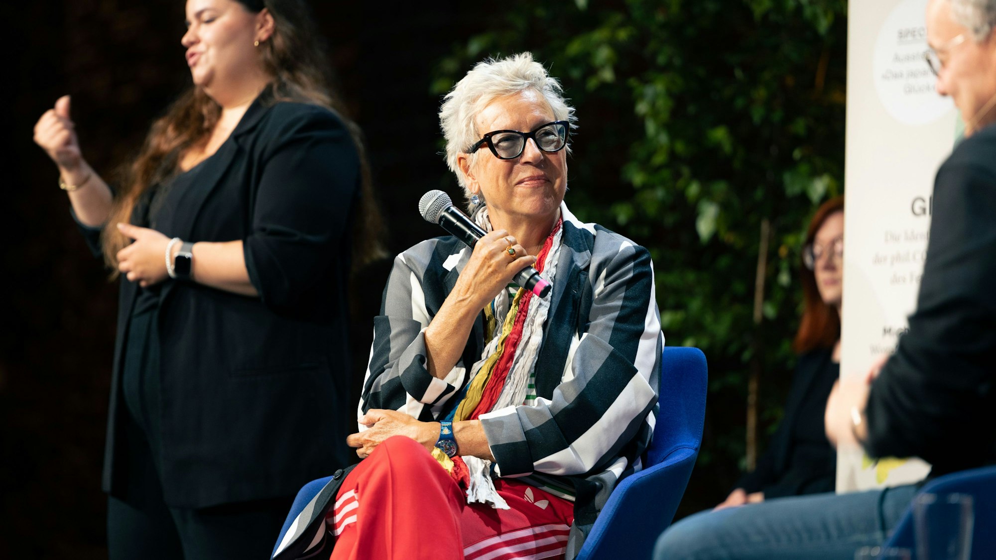 Im Vordergrund ist Doris Dörrie zu sehen, in schwarz-weiß-gestreifter Jacke und roter Jogginghose. Sie ist dem Moderator Knut Elstermann zugewandt. Im Hintergrund dolmetscht eine junge Frau das Gespräch in die Gebärdensprache.