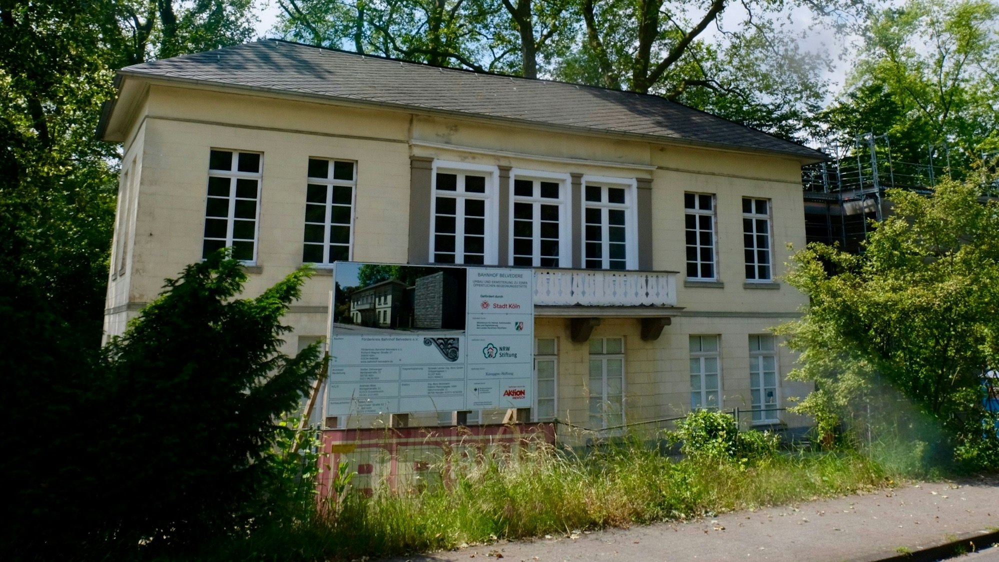 Vor der Front des ehemaligen Bahnhofs Belvedere in Müngersdorf steht ein Baustellenschild, das auf Renovierungsarbeiten hinweist. Das zweistöckige Gebäude ist zart gelb gestrichen und verfügt über sieben Fenster pro Etage, wobei sich vor drei Fenstern im ersten Geschoss ein Balkon befindet. Bäume und Sträucher säumen das Gebäude.