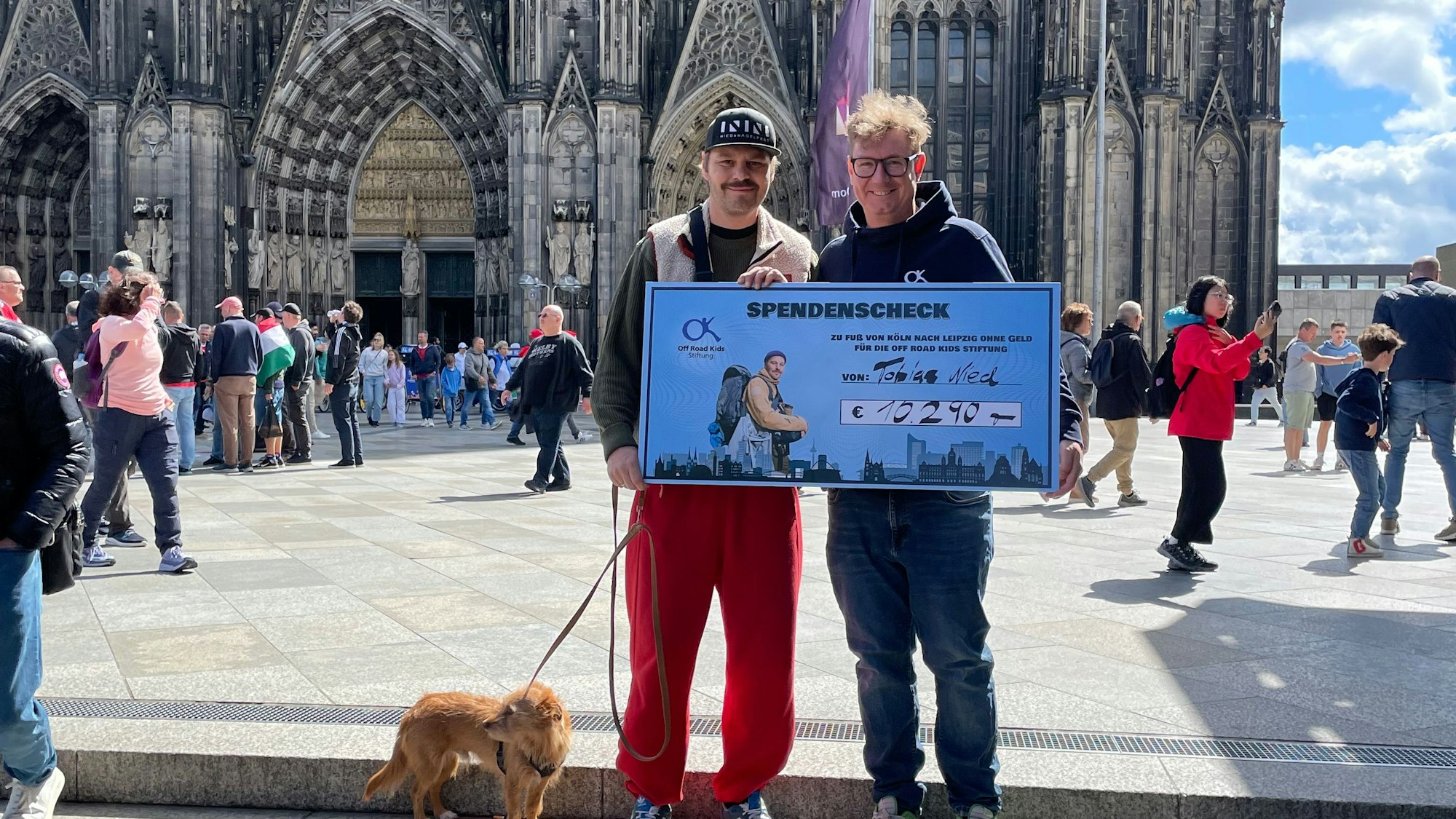 Tobias Nied (l.) mit Vetreter von Off-Roads-Kids vor dem Kölner Dom. Sie halten den Spendenscheck.