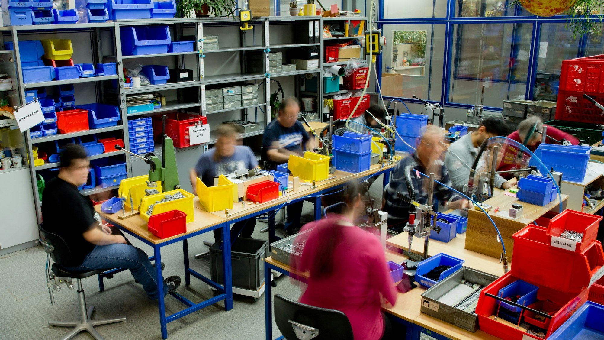 Eine Werkstatt ist zu sehen mit Tischen voller Behälter. Sieben Menschen sitzen dort und gehen einer Tätigkeit nach.