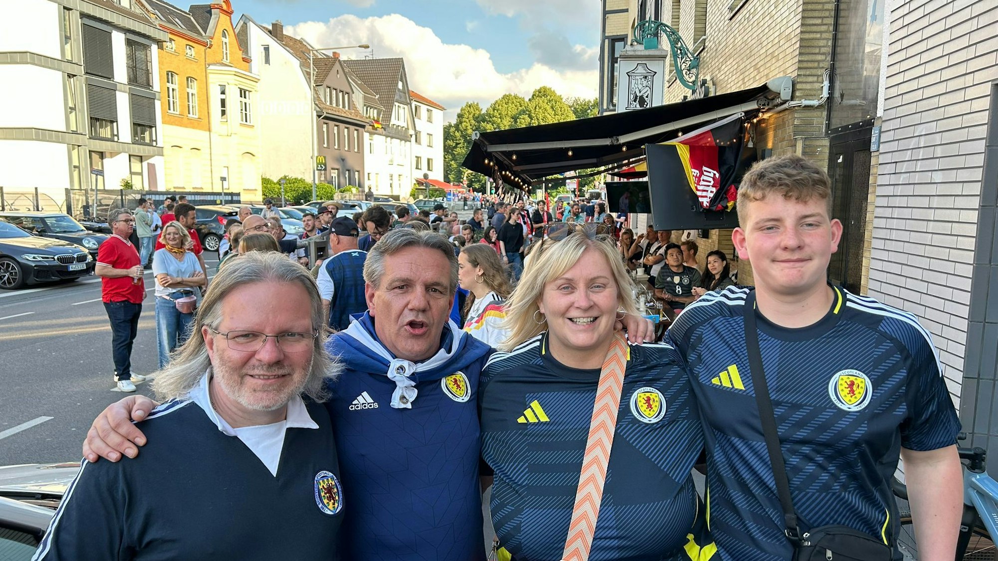 Vier schottische Fans lächeln in die Kamera. Im Hintergrund ist ein voller Außengastronomie-Bereich zu sehen.