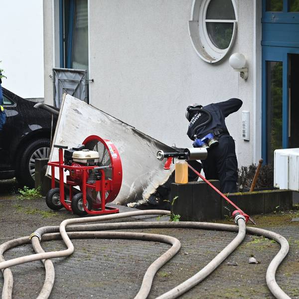 Ein Polizist steht vor der Eingangstür zu einem Mehrfamilienhaus. Zwei THW-Kräfte schauen ihm zu.