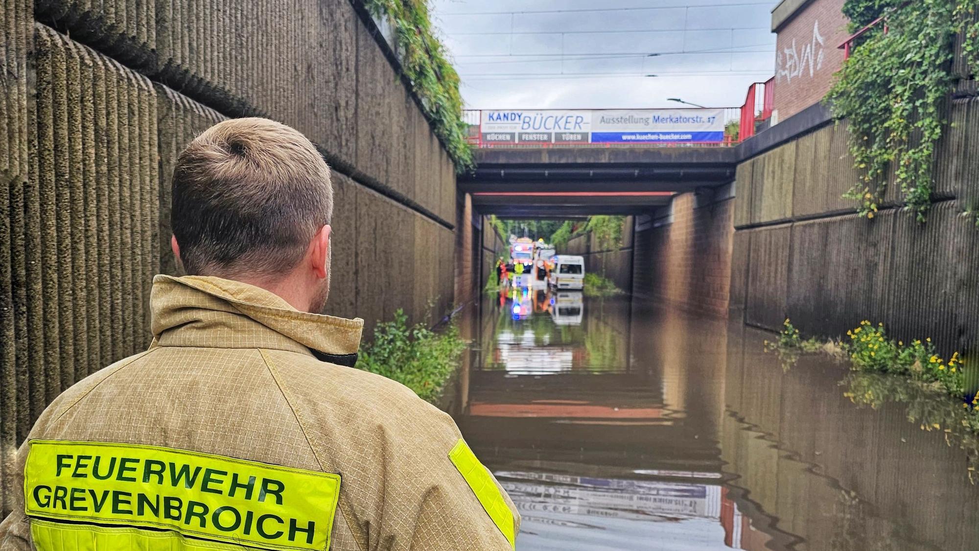 Im Vordergrund eine Einsatzkraft der Feuerwehr, im Hintergrund eine Unterführung voller Wasser, ein Transporter und weitere Fahrzeuge im Hintergrund.