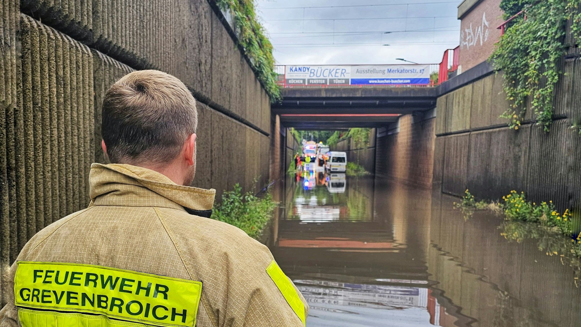 Im Vordergrund eine Einsatzkraft der Feuerwehr, im Hintergrund eine Unterführung voller Wasser, ein Transporter und weitere Fahrzeuge im Hintergrund.