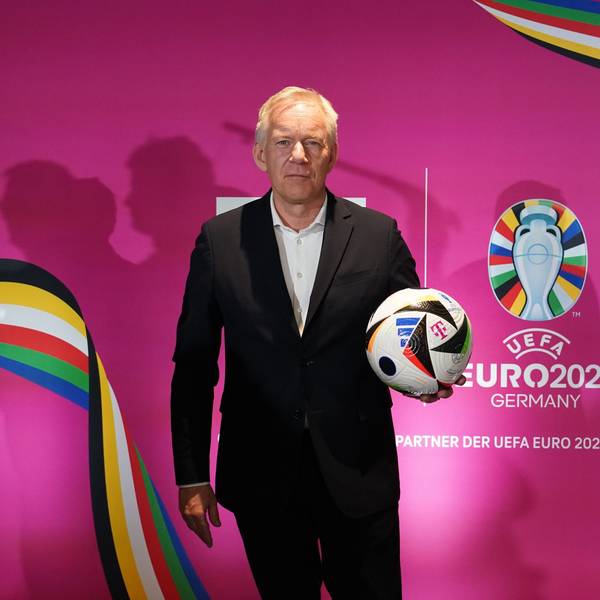 Moderator Johannes B. Kerner steht mit einem Fußball vor einer pinken Wand des Fernsehsenders MagentaTV, auf dem das Logo der Fußball-Europameisterschaft 2024 zu sehen ist