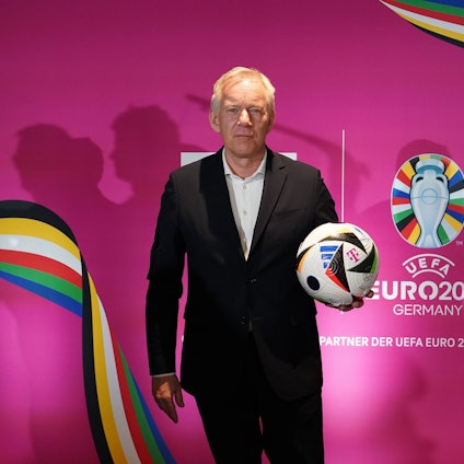 Moderator Johannes B. Kerner steht mit einem Fußball vor einer pinken Wand des Fernsehsenders MagentaTV, auf dem das Logo der Fußball-Europameisterschaft 2024 zu sehen ist