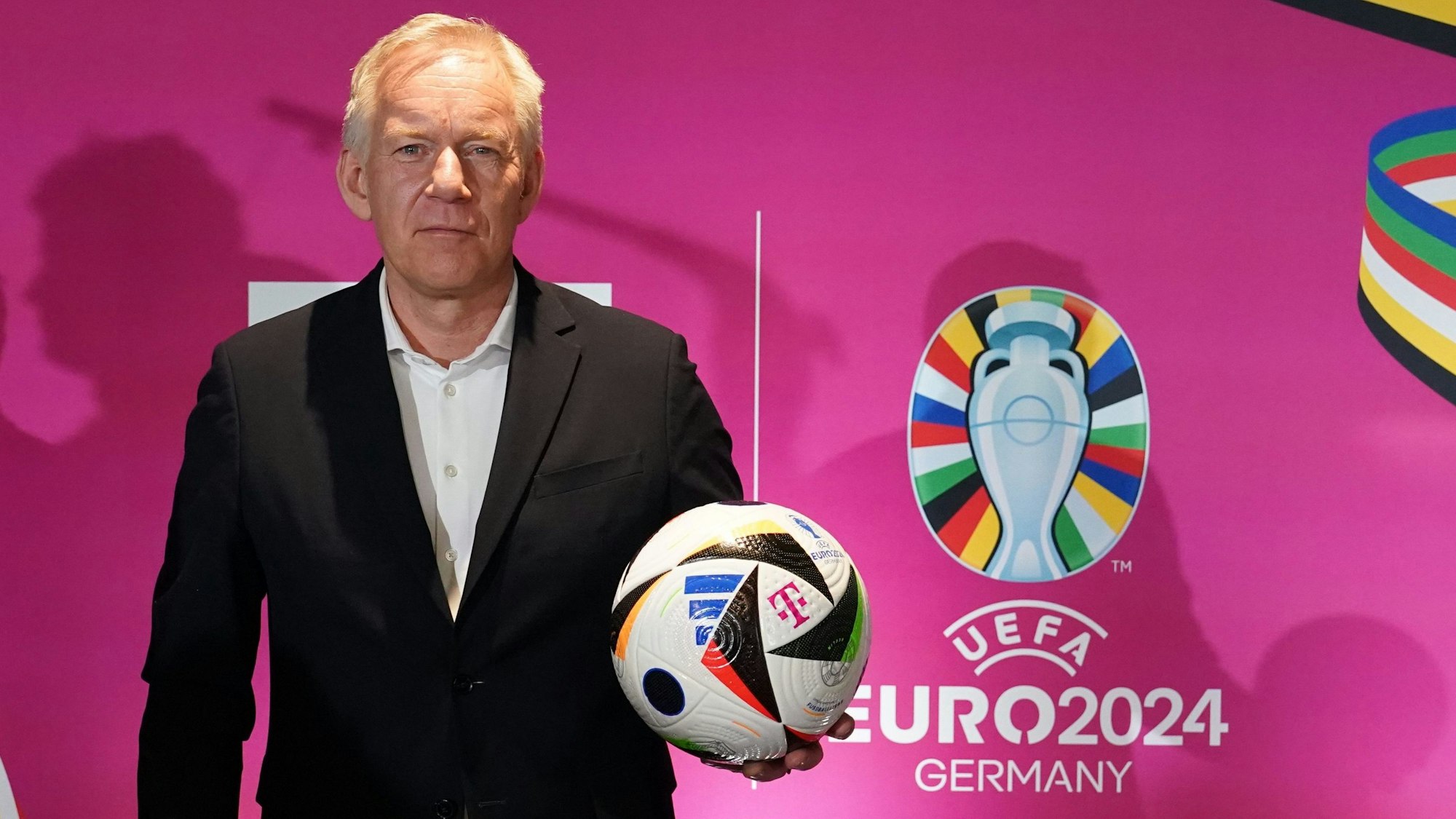 Moderator Johannes B. Kerner steht mit einem Fußball vor einer pinken Wand des Fernsehsenders MagentaTV, auf dem das Logo der Fußball-Europameisterschaft 2024 zu sehen ist